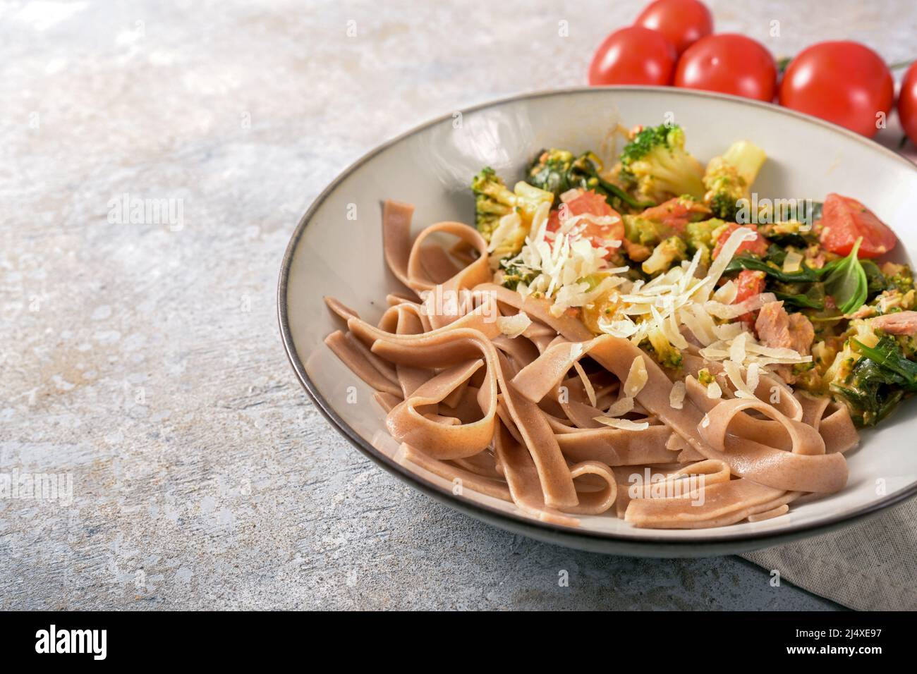 Piatto sano di tagliatelle a grani interi con verdure come broccoli, pomodori e spinaci, servito con parmigiano in un piatto su un rustico grigio Foto Stock