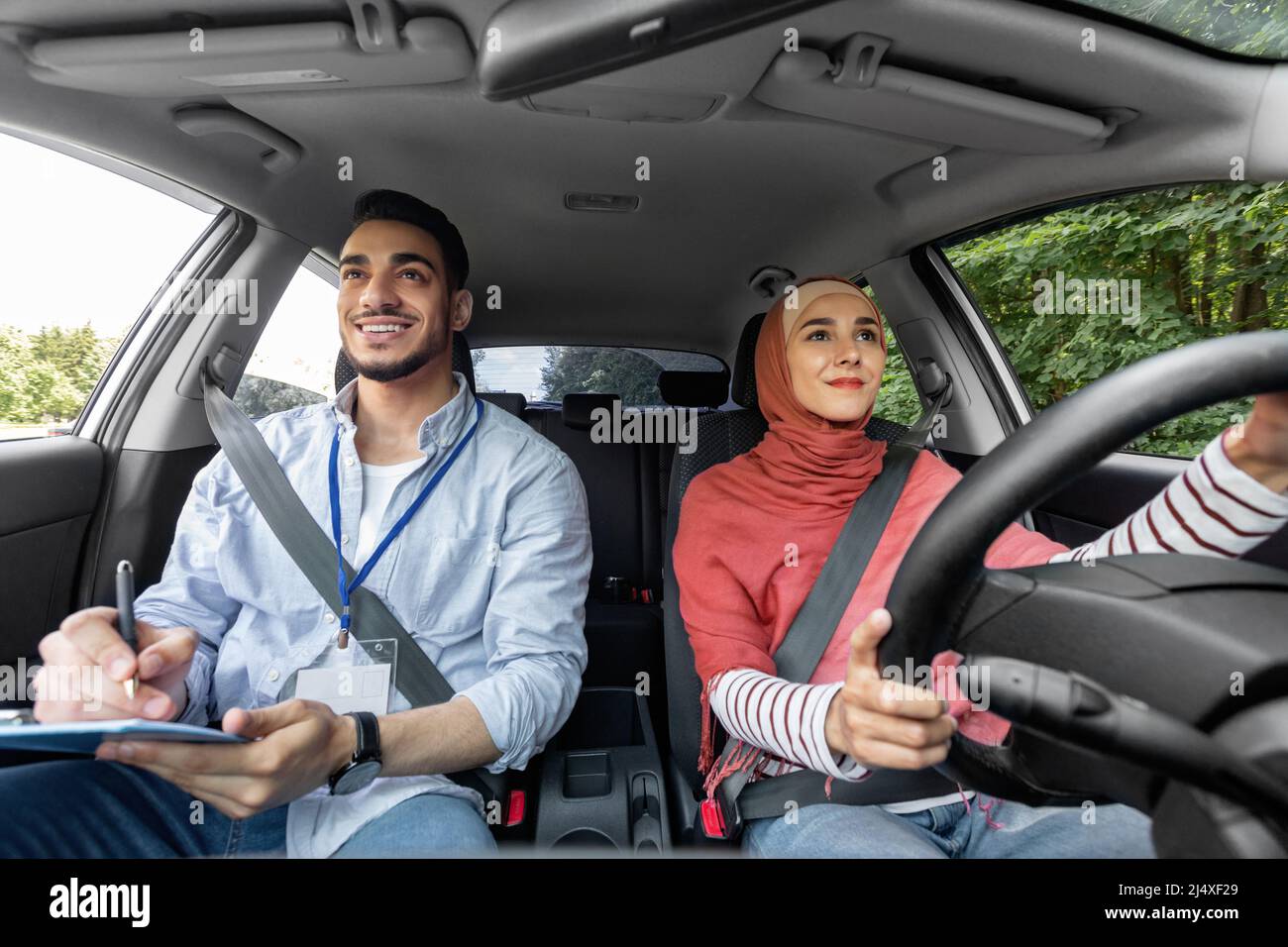 Sorridente donna araba millenaria in hijab che prende la prova di guida con l'uomo mentore, signora al volante guida in auto Foto Stock