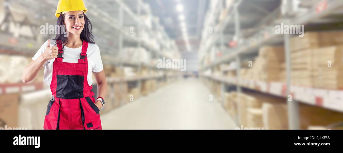 Un addetto al magazzino punta il dito verso l'alto. Sorridente giovane donna in abiti da lavoro di fronte ad un enorme magazzino. Foto Stock