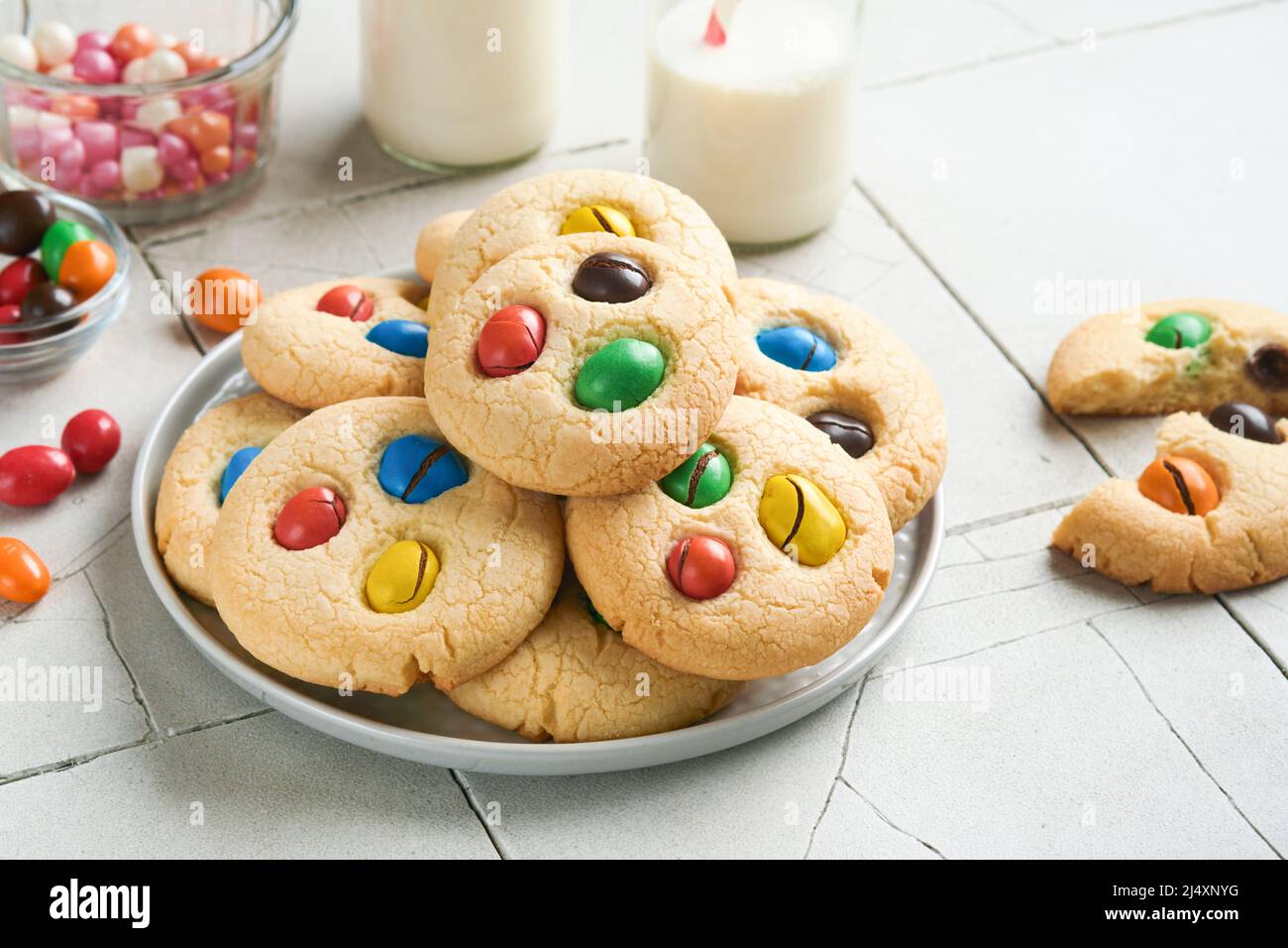 Biscotti fatti in casa con caramelle colorate al cioccolato e latte. Accatastate biscotti frollini con caramelle multicolore sul piatto con una bottiglia di latte accesa Foto Stock