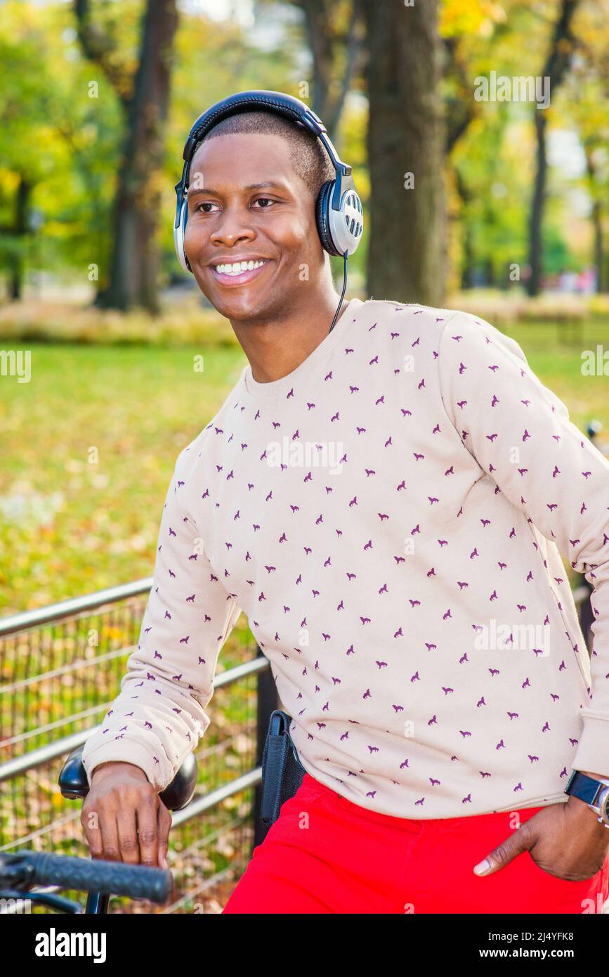 Amore uomo Musica. Vestirsi in maglione senza collari a motivi geometrici,  pantaloni rossi, indossare cuffie, un giovane ragazzo nero si inclina su  una bicicletta sul parco, sorridendo Foto stock - Alamy