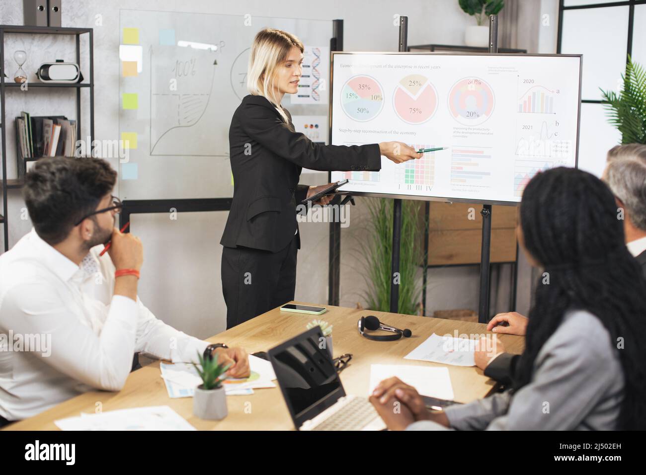 Bella e motivata giovane donna d'affari bionda intelligente che dimostra l'infografia sul bordo digitale di fronte al suo concentrato di business multietnico team, ascoltare e prendere appunti. Foto Stock