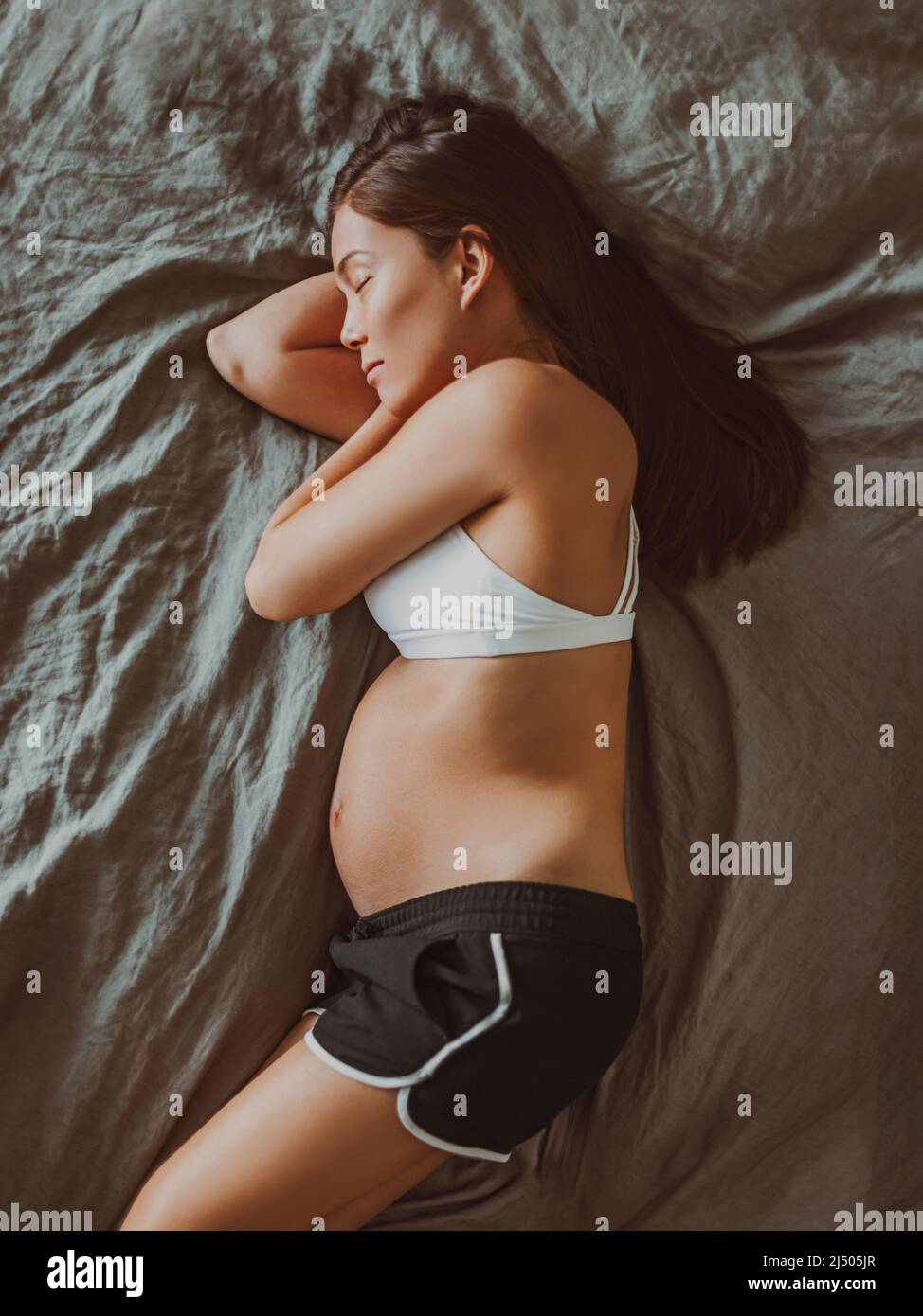 Donna incinta che dorme vista dall'alto del ventre di gravidanza durante il primo trimestre. Ragazza asiatica in pigiama sdraiata sul suo fianco per dormire a letto. Ritaglio verticale Foto Stock