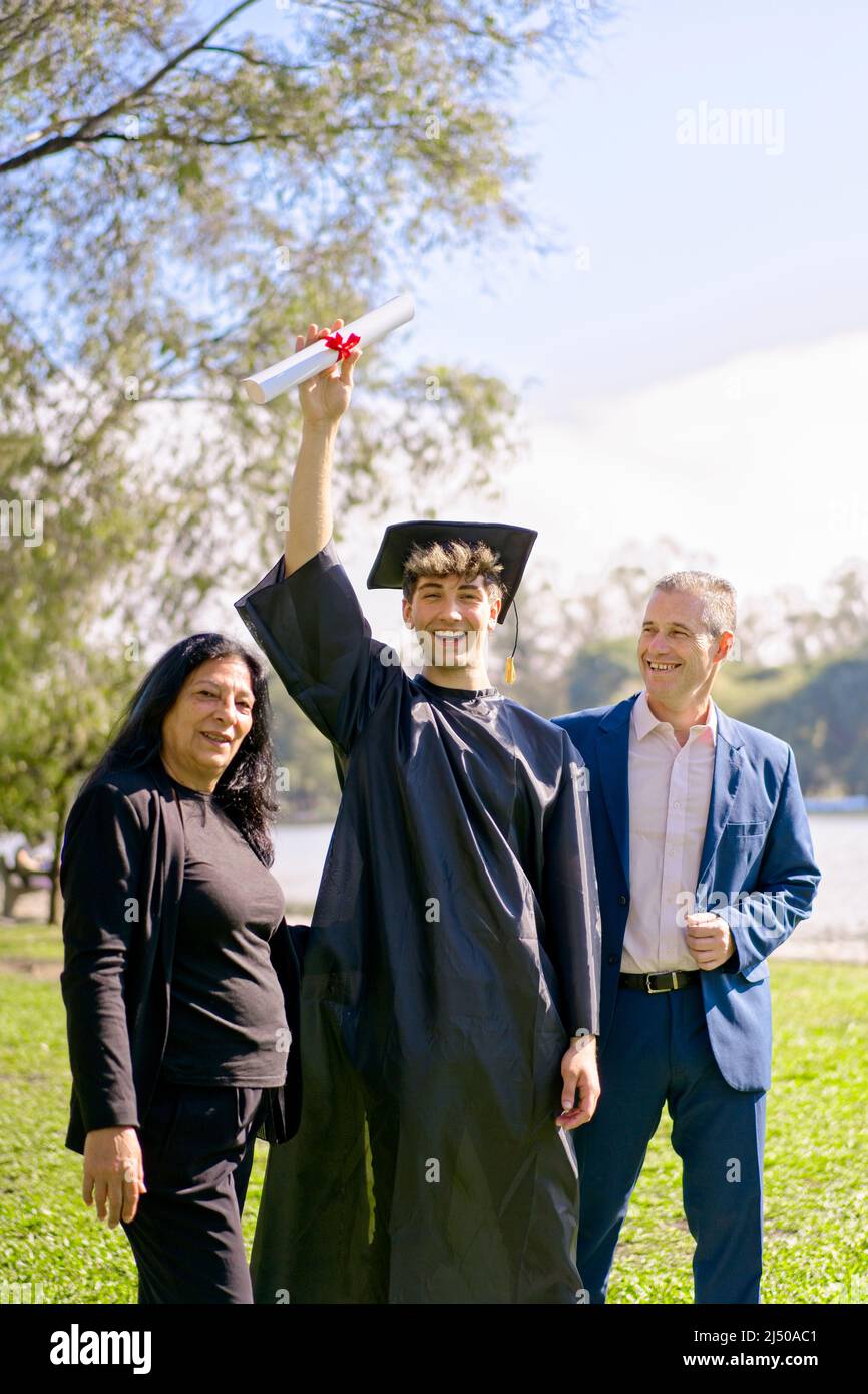 Giovane si è laureato di recente, vestito di cappello e abito, con la sua laurea in mano, festeggiando con la sua famiglia multietnica nel campus universitario. Ve Foto Stock