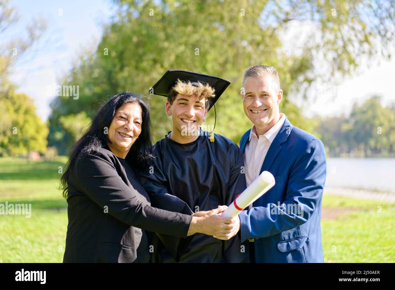 Giovane si è laureato di recente, vestito di cappello e abito, con la sua laurea in mani, festeggiando con la sua famiglia multietnica nel campus universitario. V Foto Stock