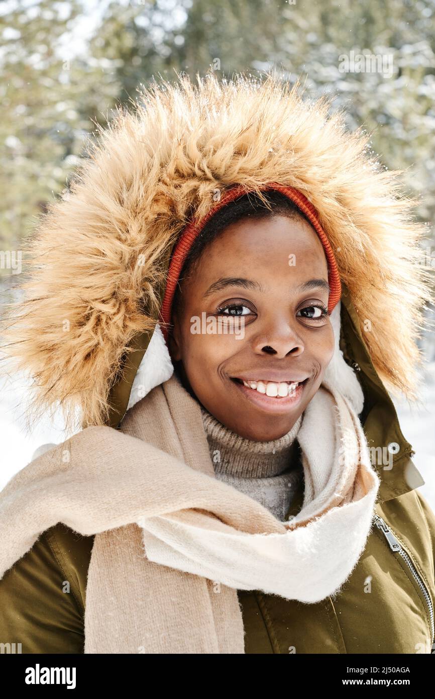 Allegra giovane donna nera in calda giacca invernale con cappuccio che guarda la macchina fotografica mentre si trova in mezzo al bosco in una giornata gelida Foto Stock