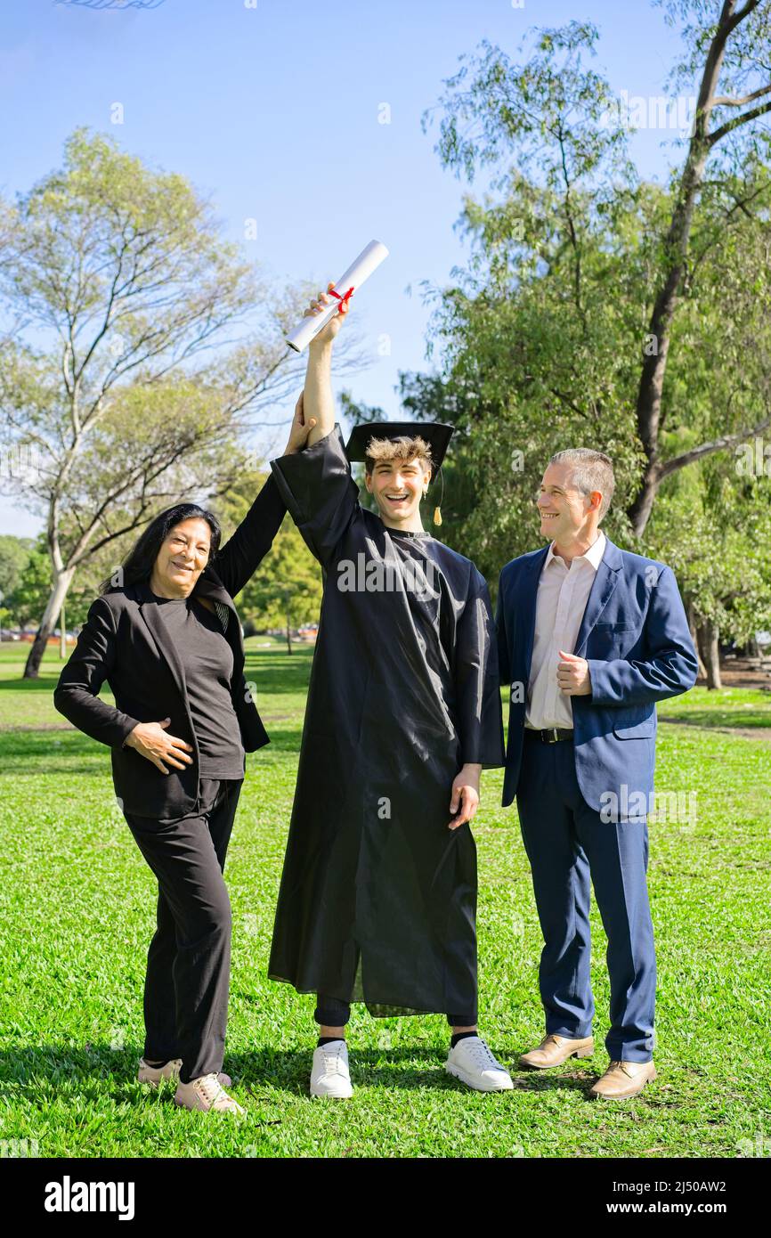 Giovane si è laureato di recente, vestito di cappello e abito, con la sua laurea in mano, festeggiando con la sua famiglia multietnica nel campus universitario. Ve Foto Stock