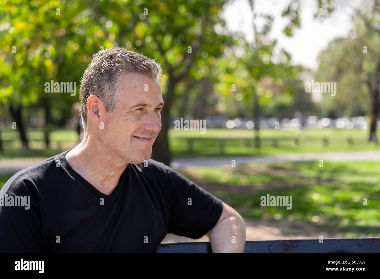 Primo piano ritratto di un uomo maturo con capelli grigi indossando una t-shirt nera seduta su una panca del parco guardando lateralmente Foto Stock