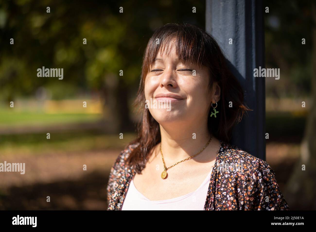 Ritratto di una giovane donna latina in un parco appoggiato su un lampione, godendo i raggi del sole sulla pelle, con gli occhi chiusi Foto Stock