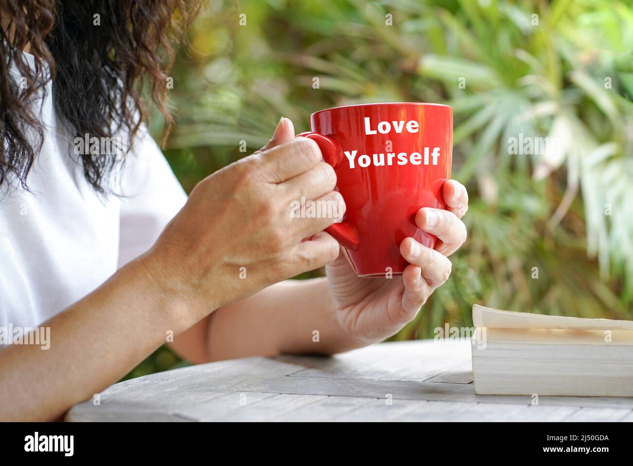 Donna che tiene una tazza rossa con il testo Love Yourself, e un libro sul tavolo. Esterno con sfondo di palme. Foto Stock