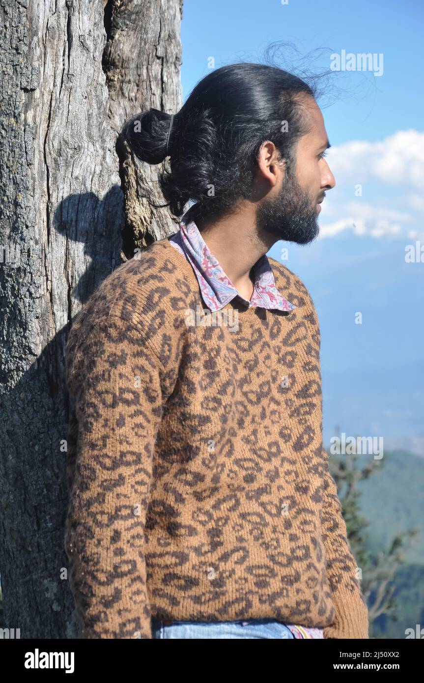 Un buon uomo giovane con capelli bun guardare lontano mentre in piedi contro albero nella natura Foto Stock