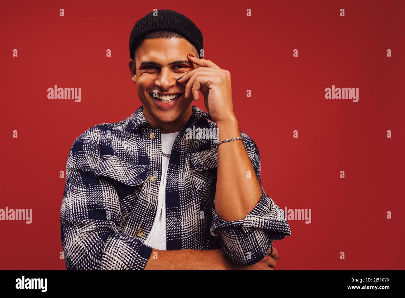 Giovane uomo di stile sorridente allegro in uno studio. Felice giovane uomo in piedi da solo su sfondo rosso. Giovane uomo alla moda che indossa smalto per unghie A. Foto Stock