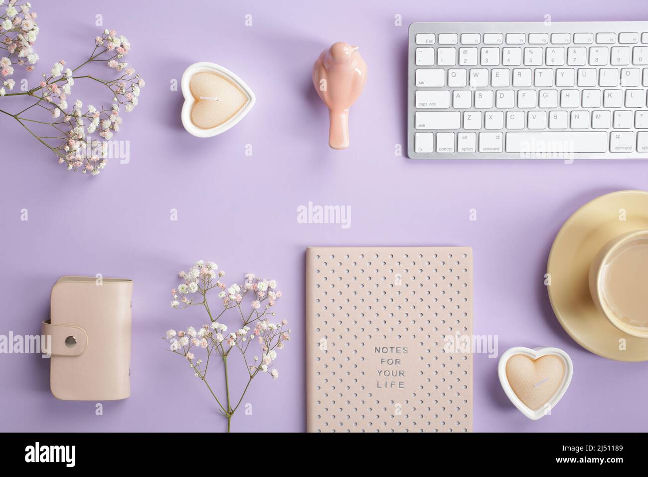 Accogliente tavolo da ufficio con tastiera, notebook, tazza da caffè, accessori e fiori di gypsophila su sfondo viola pastello. Elegante lavoro femminile Foto Stock