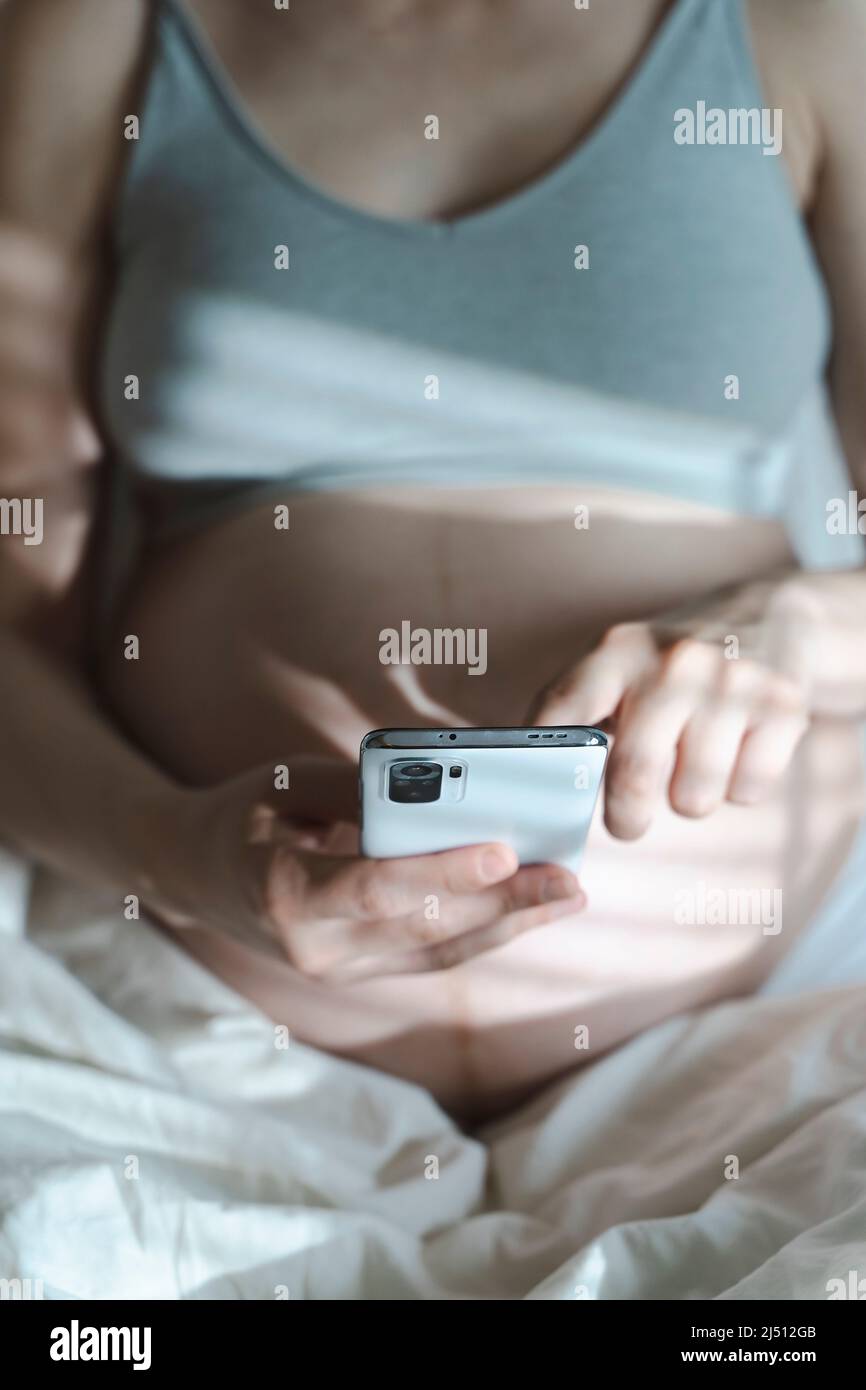 Una donna incinta usa uno smartphone mentre si rilassa nel suo letto di casa. Internet, shopping, comunicazione durante la gravidanza e prima della nascita di un bambino. Salute delle donne e dei neonati. Foto di alta qualità Foto Stock