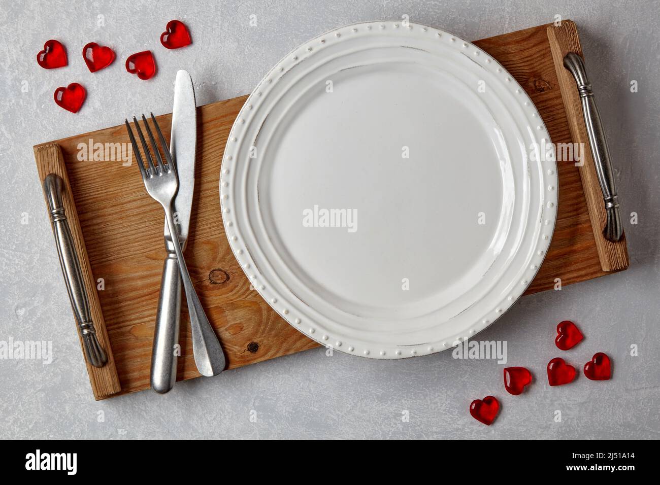 Piatto bianco vuoto con posate su vassoio di legno su un tavolo di cemento grigio chiaro decorato con cuori di caramello rosso Foto Stock
