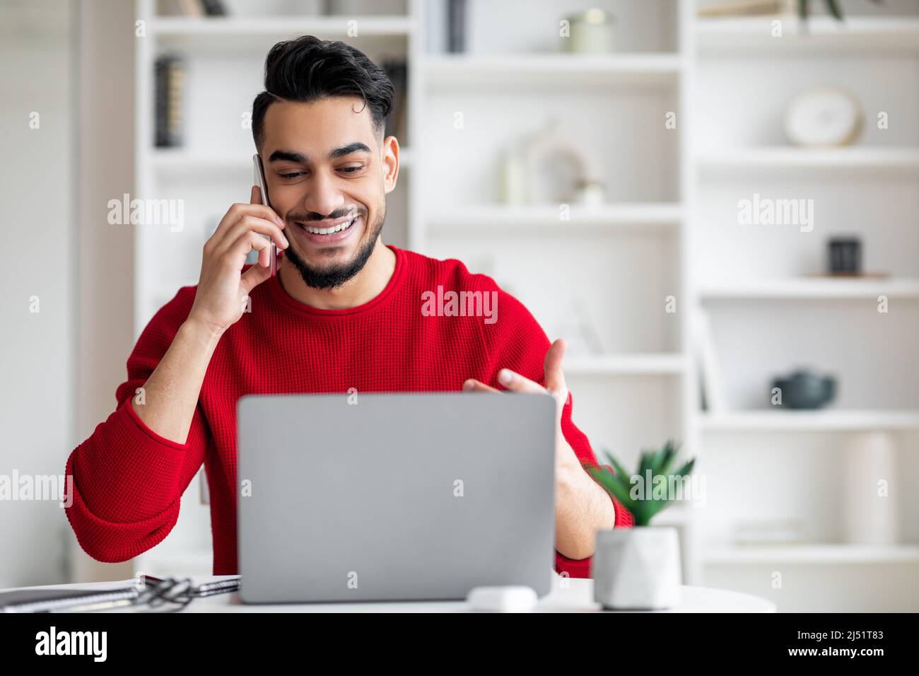 Sorridente maschio arabo millenario con barba in abiti rossi parla per telefono e guarda il computer portatile Foto Stock