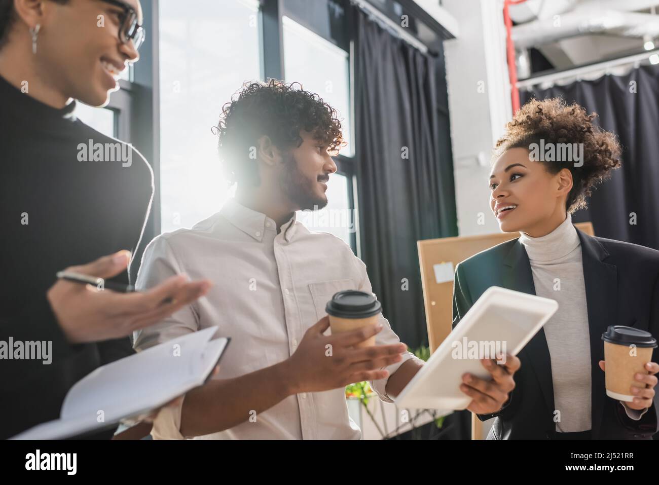 Allegri uomini d'affari multietnici con caffè da andare e tablet digitale che parlano in ufficio Foto Stock