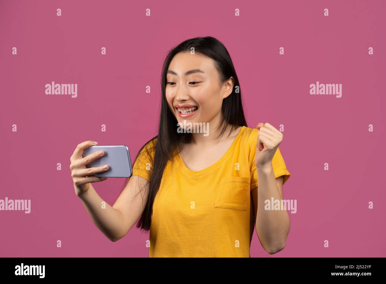 Sì, grandi notizie. Signora coreana molto gioiosa che celebra la vittoria, tenendo il cellulare e scuotendo pugno clenched, sfondo violetto Foto Stock