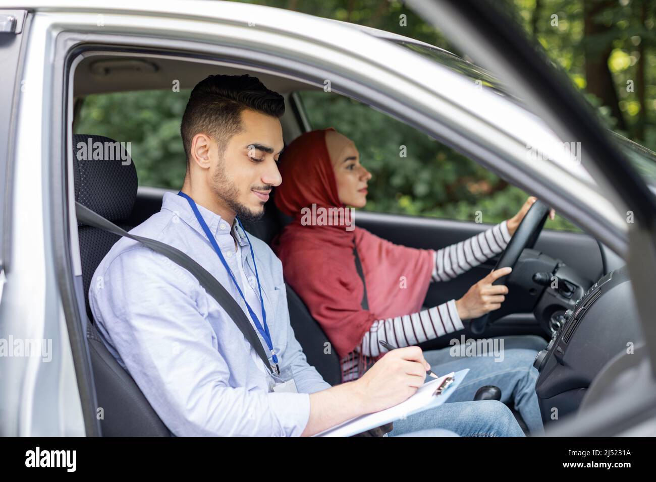 Uomo arabo serio millenario che prende test di guida da donna in hijab, signora al volante guida auto, profilo Foto Stock