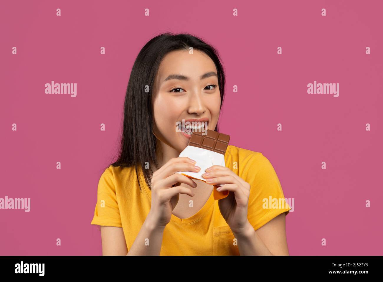 Giovane donna coreana che mangia un gustoso bar al cioccolato, gustando un dolce yummy su sfondo viola studio Foto Stock