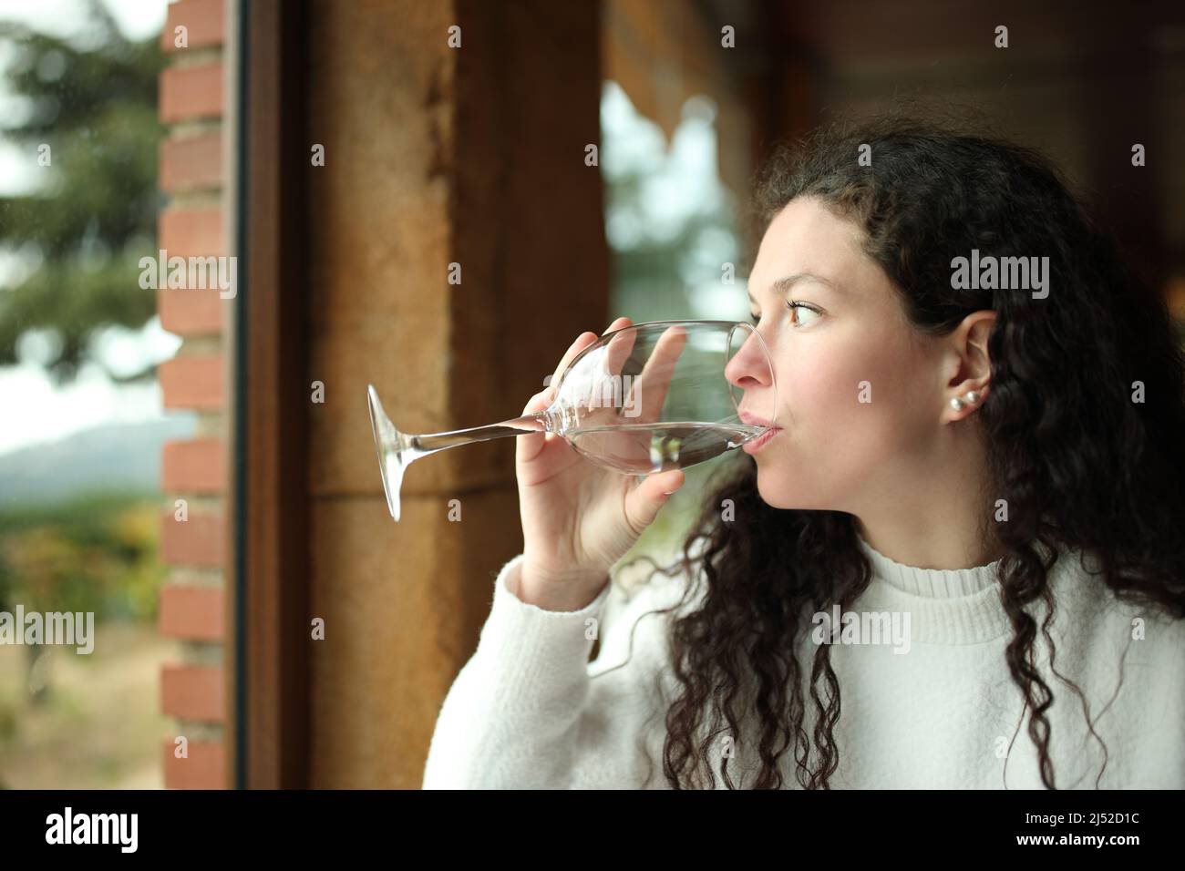 Donna distratta che beve acqua guardando attraverso una finestra in una casa Foto Stock
