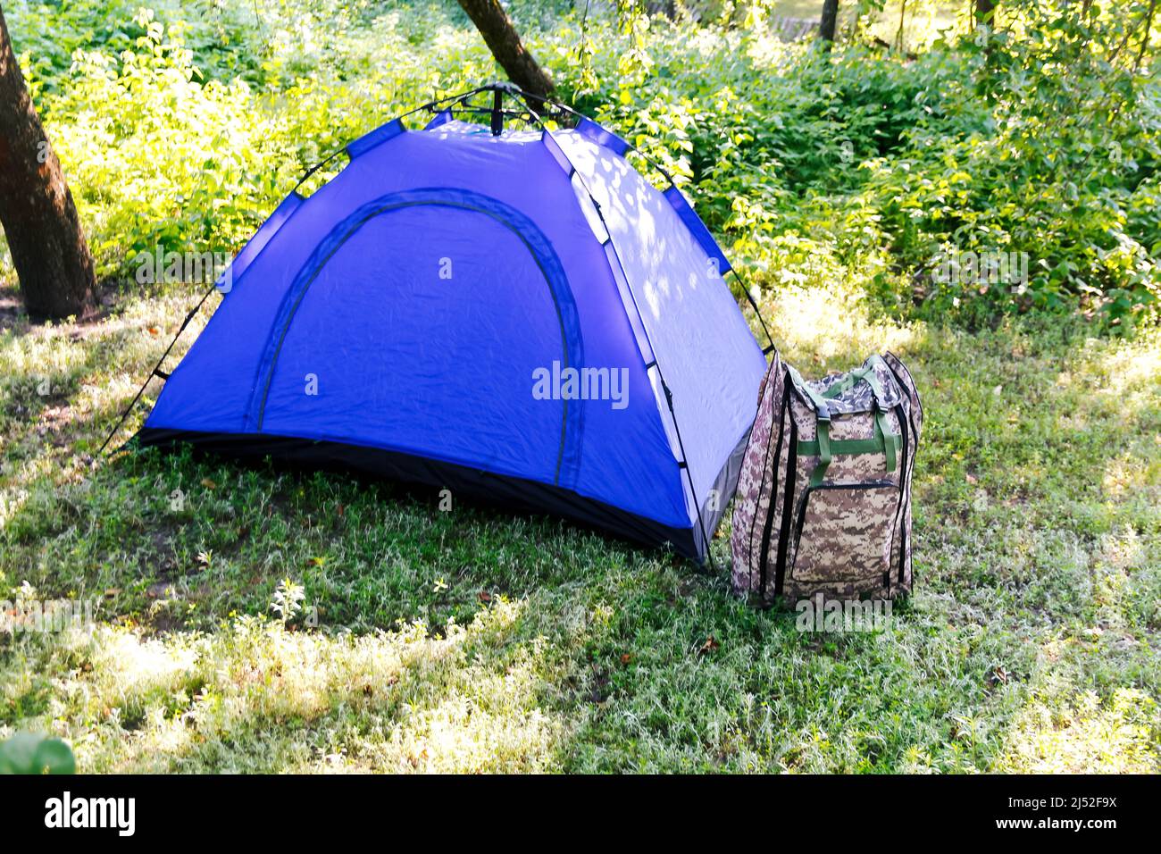 Sfocatura blu tenda turistica aperta in piedi su sfondo verde natura. Zaino militare. Concetto di turismo. Vacanze estive nella foresta, campeggio. Fuori fuoco Foto Stock