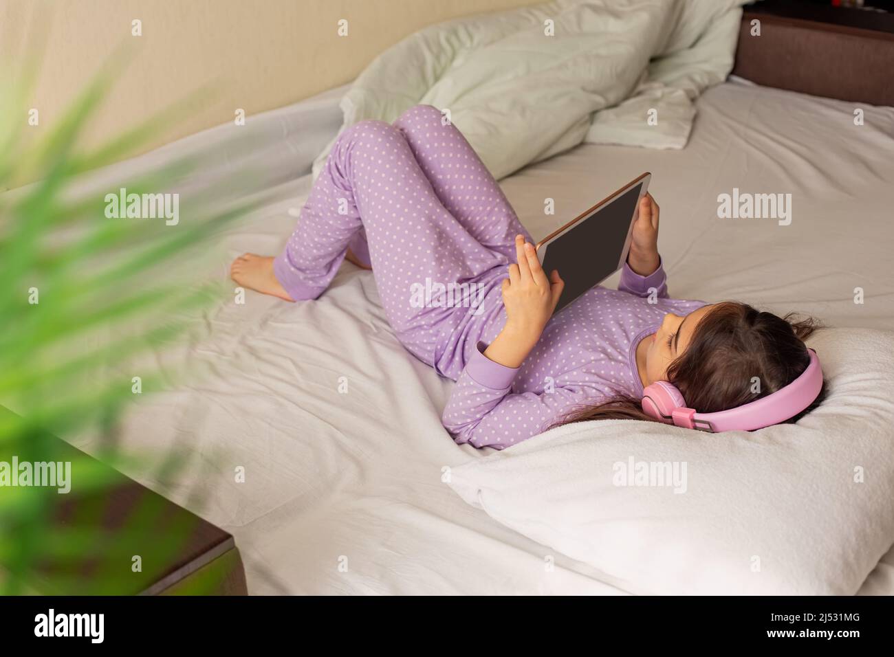 Una ragazza in pigiama viola, si trova su un letto bianco sulla schiena, tiene in mano un tablet digitale rosa. Foto Stock
