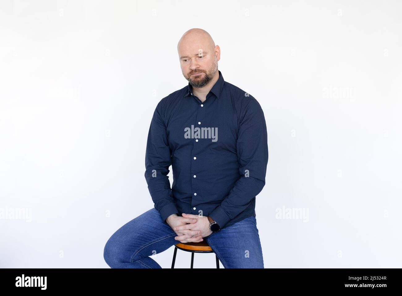Ritratto di uomo di mezza età in camicia blu, jeans seduti su sgabello, dita interlacciate su sfondo bianco. Foto Stock
