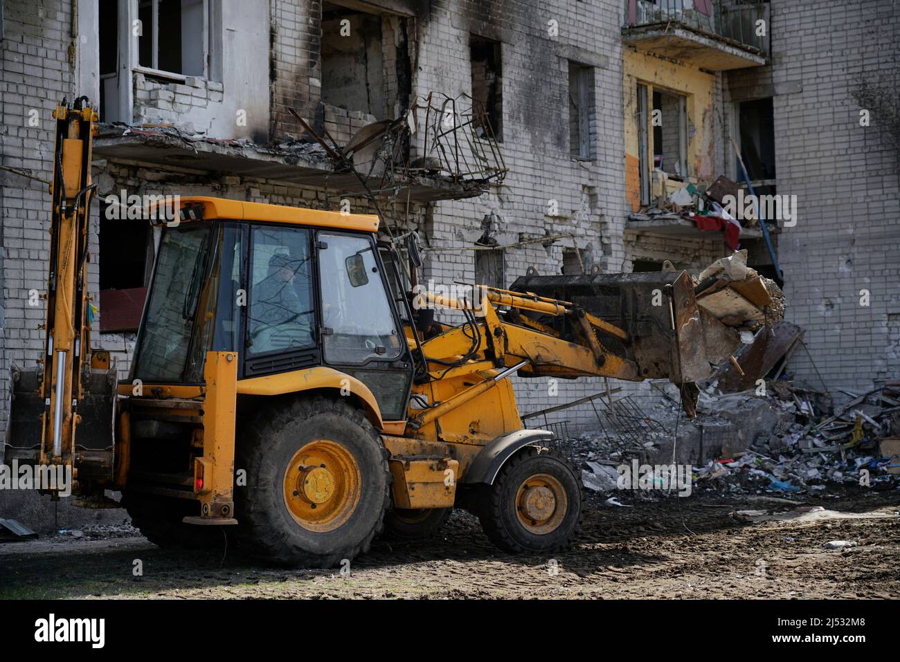 Un trattore elimina i rifiuti vicino a una casa di città distrutta. Un grande edificio residenziale senza finestra fu danneggiato da un'esplosione durante la guerra a Ukra Foto Stock