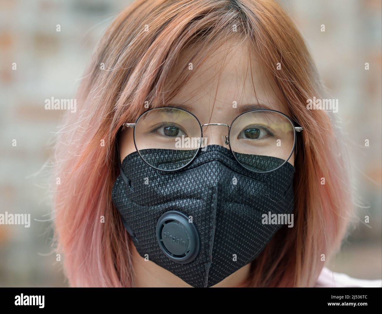 Giovane donna vietnamita graziosa con i capelli tinti e gli occhiali indossa una maschera protettiva nera con valvola di respirazione durante la pandemia di coronavirus. Foto Stock