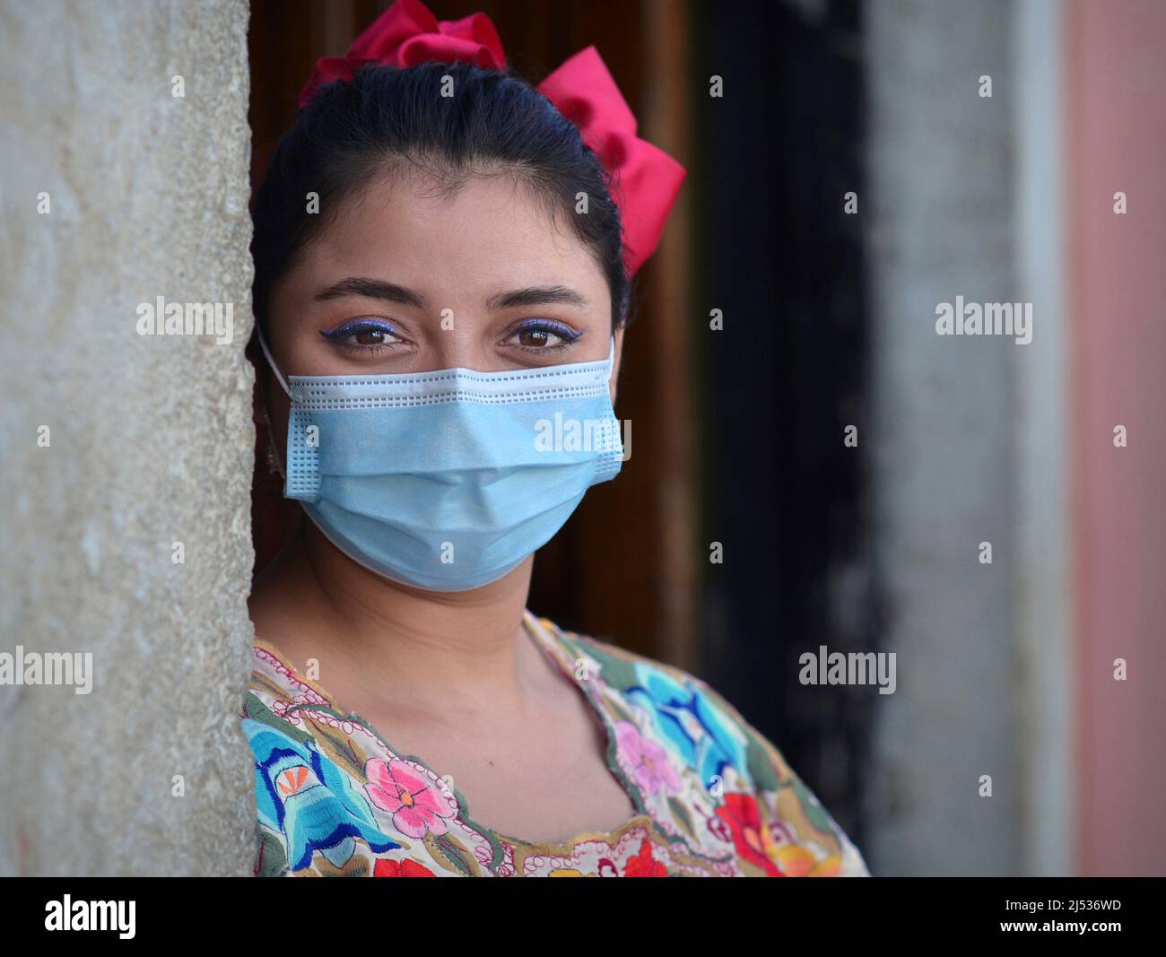 La giovane donna messicana positiva con occhi sorridenti e il trucco dell'occhio blu indossa un vestito tradizionale Maya Yucatan colorato e una maschera chirurgica blu chiaro. Foto Stock