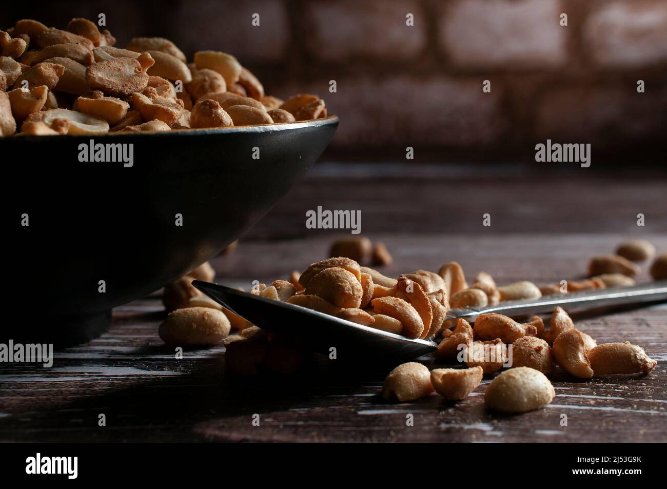 Arachidi arrosto su una ciotola nera e su un cucchiaio su un rustico tavolo di legno con una parete di mattoni sullo sfondo Foto Stock