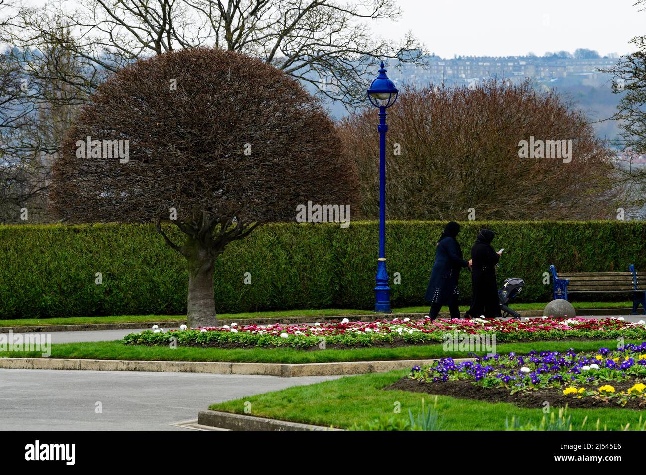 Servizi municipali urbani panoramici (camminando sul sentiero da piante da letto luminose, bordi puliti e siepe, sedile a panca) - Lister Park, Bradford, Inghilterra Regno Unito. Foto Stock