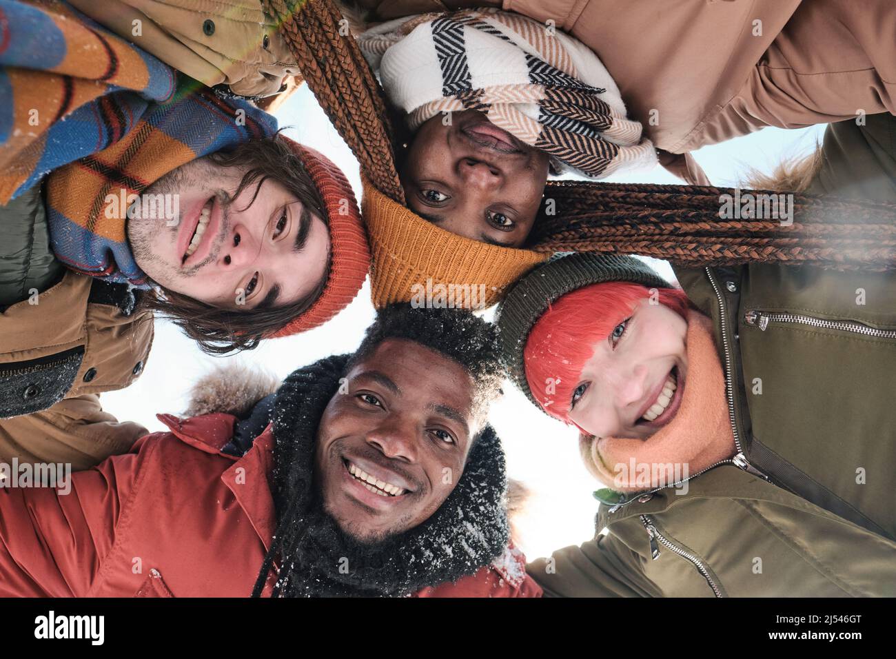 Da sotto vista di giovani amici afroamericani e caucasici che trascorrono una giornata invernale nevosa insieme sorridendo a macchina fotografica Foto Stock