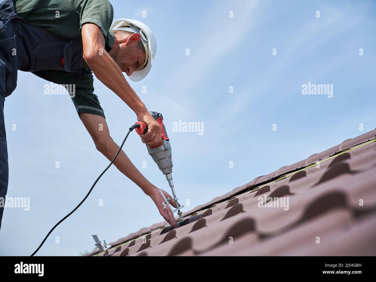 Lavoratore prepearing per l'installazione di pannelli solari fotovoltaici sul tetto della casa. Man installer che lavora con cacciavite elettrico, indossando uniforme e casco, su sfondo blu cielo. Foto Stock