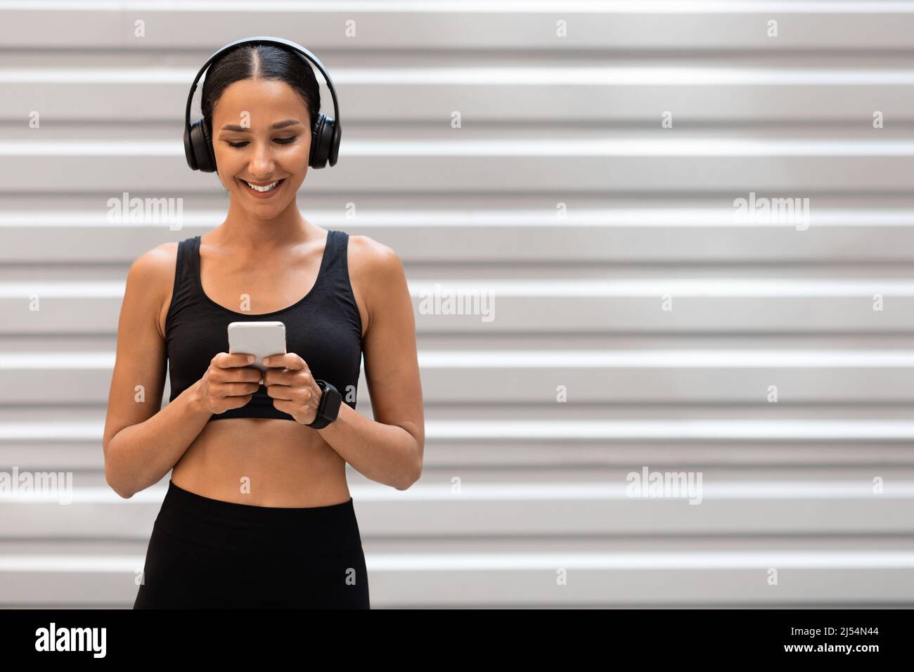 Sorridente giovane atleta araba in abbigliamento sportivo con cuffie che chiacchierano sul telefono su sfondo grigio parete Foto Stock
