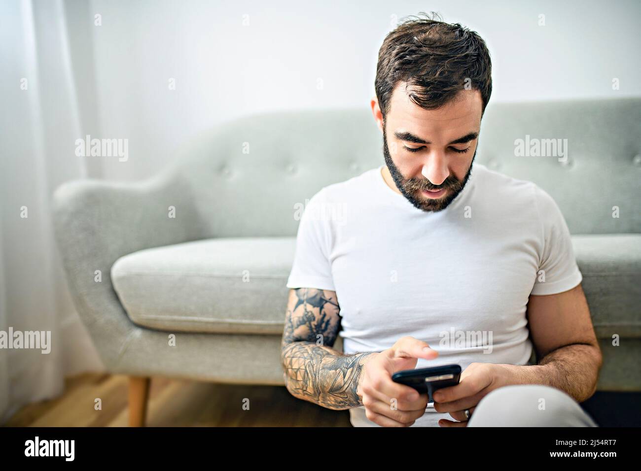 ritratto di giovane uomo casual con barba e tatuaggio e camicia bianca seduta sul divano a casa con cellulare Foto Stock