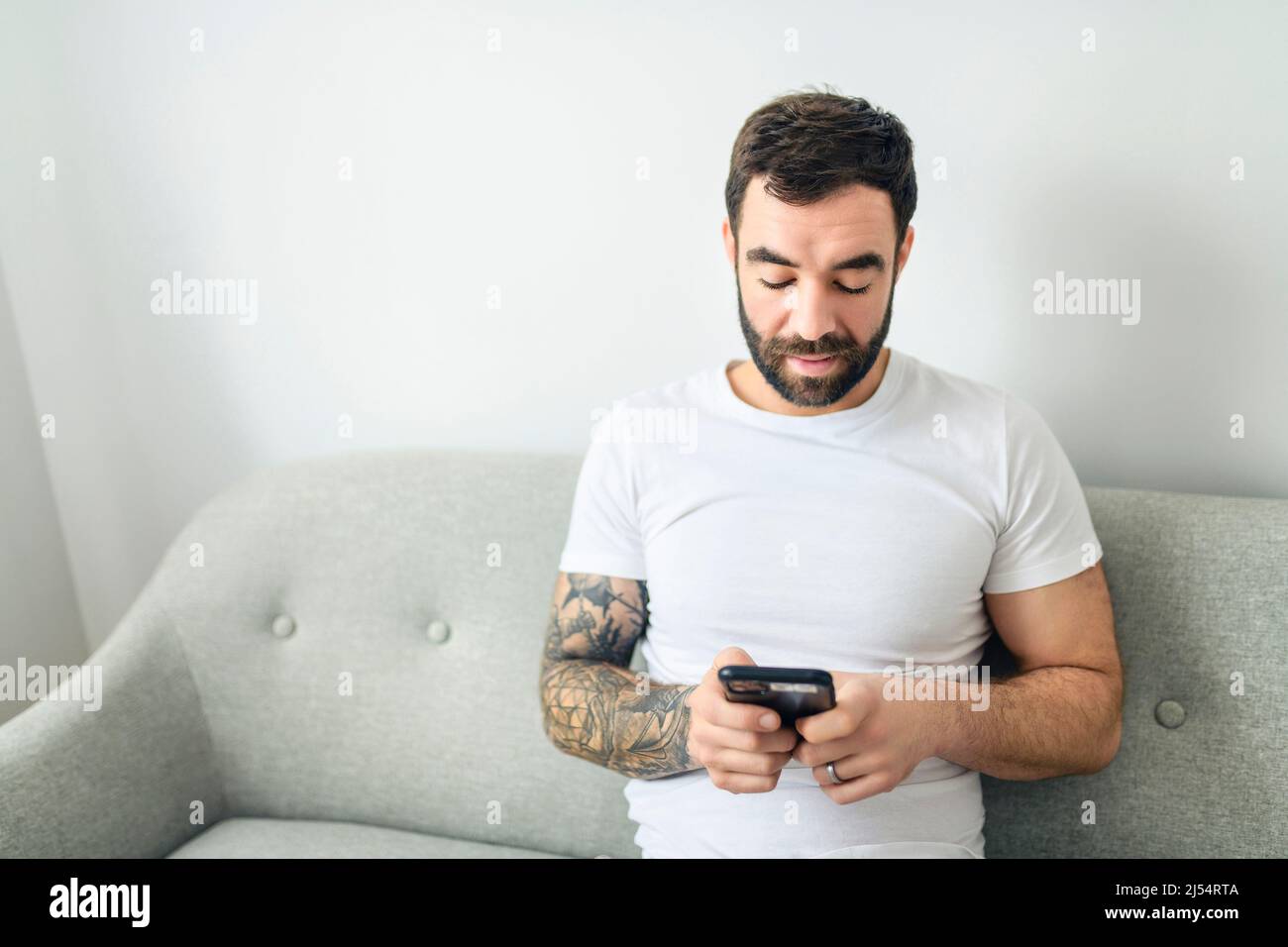 ritratto di giovane uomo casual con barba e tatuaggio e camicia bianca seduta sul divano a casa con cellulare Foto Stock