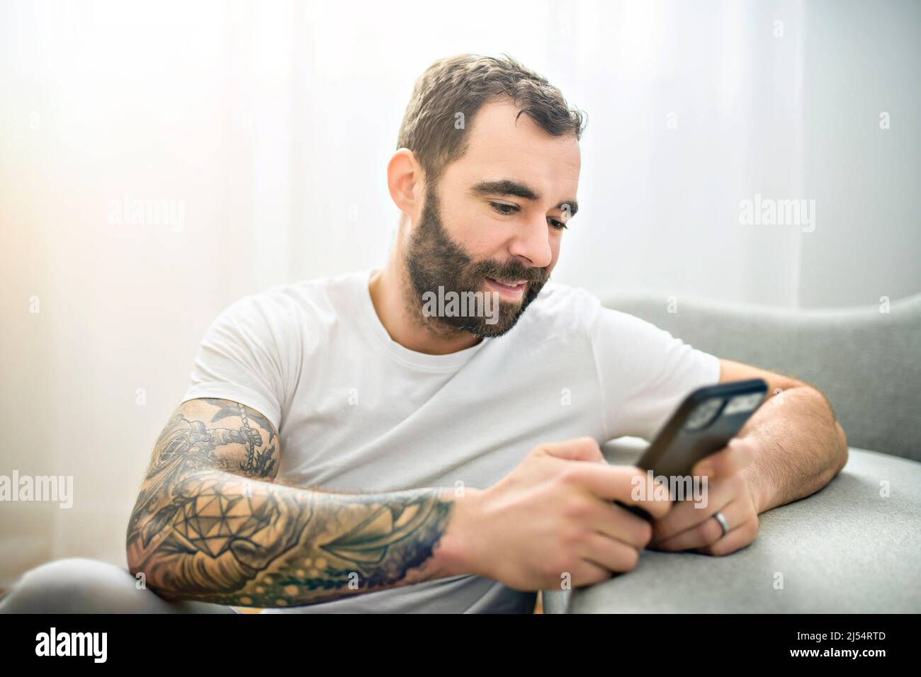 ritratto di giovane uomo casual con barba e tatuaggio e camicia bianca seduta sul divano a casa con cellulare Foto Stock