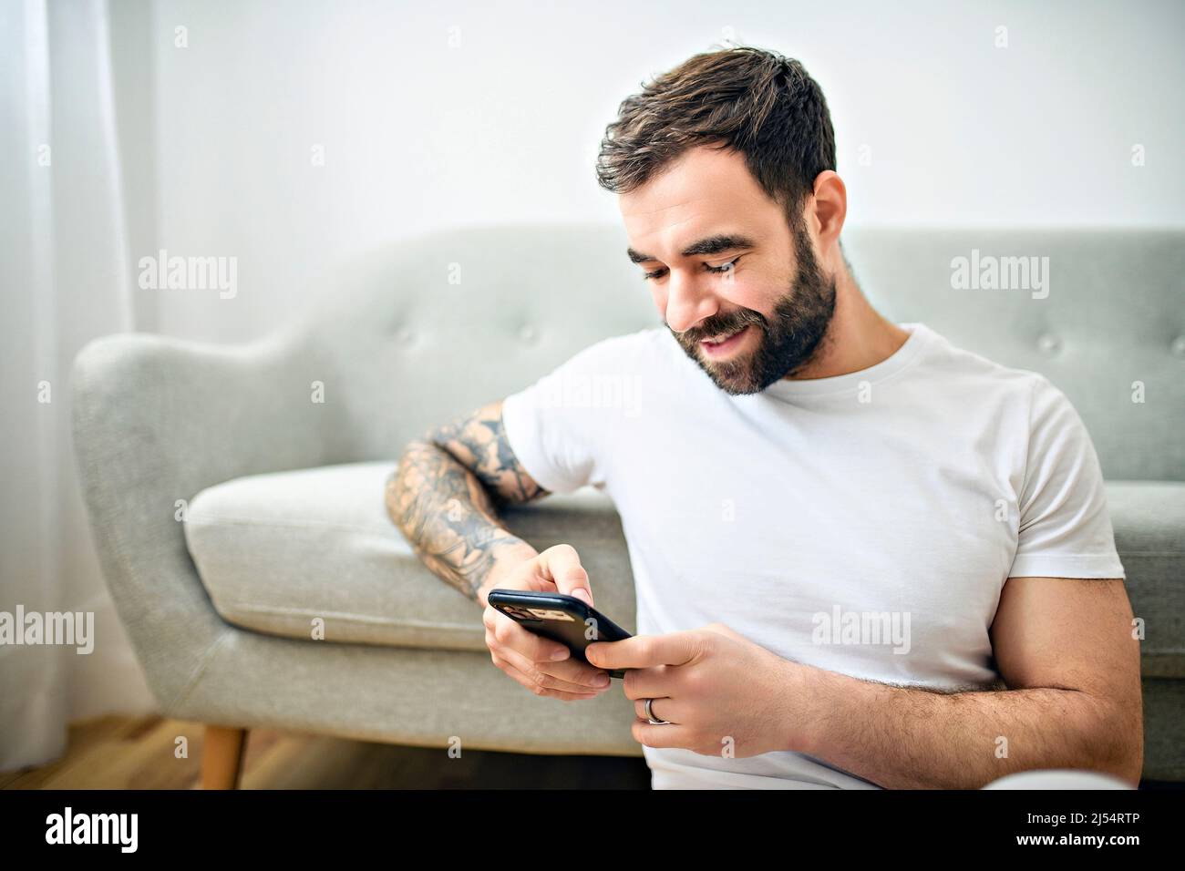 ritratto di giovane uomo casual con barba e tatuaggio e camicia bianca seduta sul divano a casa con cellulare Foto Stock