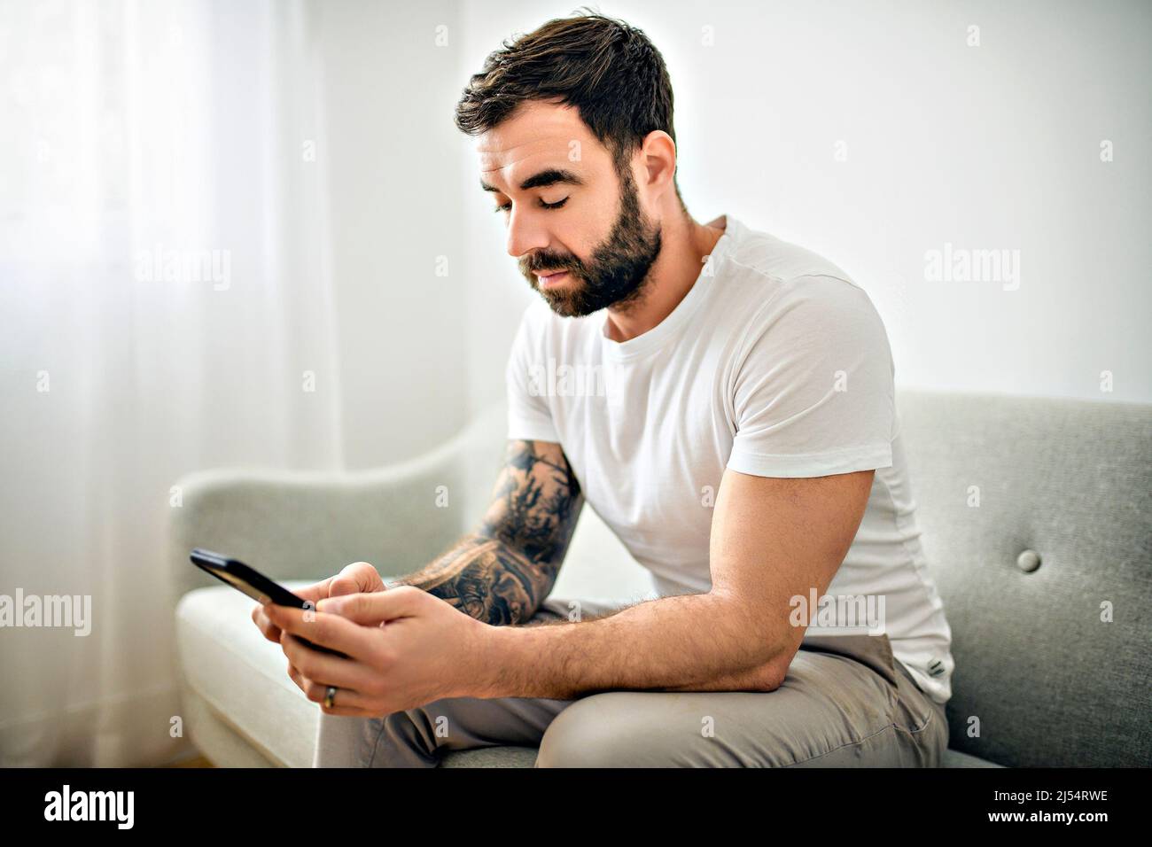 ritratto di giovane uomo casual con barba e tatuaggio e camicia bianca seduta sul divano a casa con cellulare Foto Stock