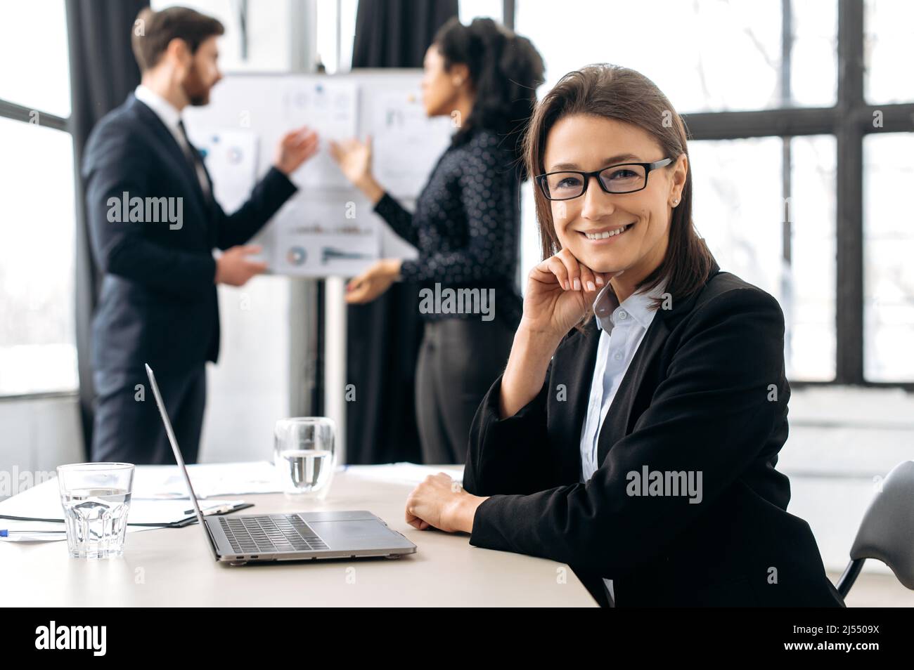 Donna da lavoro caucasica intelligente con occhiali, seduta alla scrivania alla conferenza, vicino a un computer portatile. Bella e elegante dipendente femminile in riunione briefing, guardando la fotocamera, sorridente amichevole Foto Stock