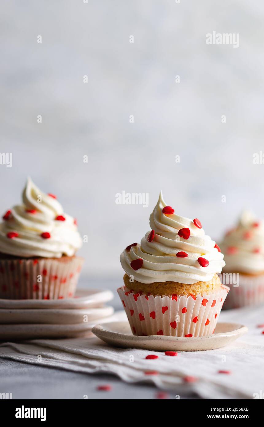 Muffin alla vaniglia con glassa alla crema spolverata con cuori di zucchero rosso sui piatti e un panno bianco, su sfondo grigio con spazio per la copia. Foto Stock