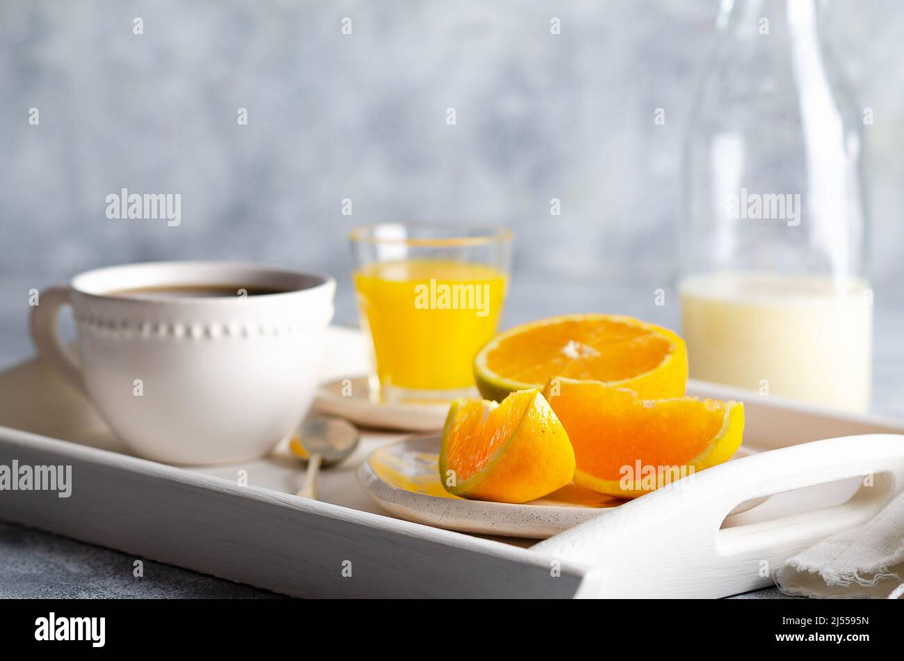 Una bottiglia di latte, una tazza di caffè, succo d'arancia e pezzi d'arancia, in un vassoio bianco e su sfondo grigio. Foto Stock