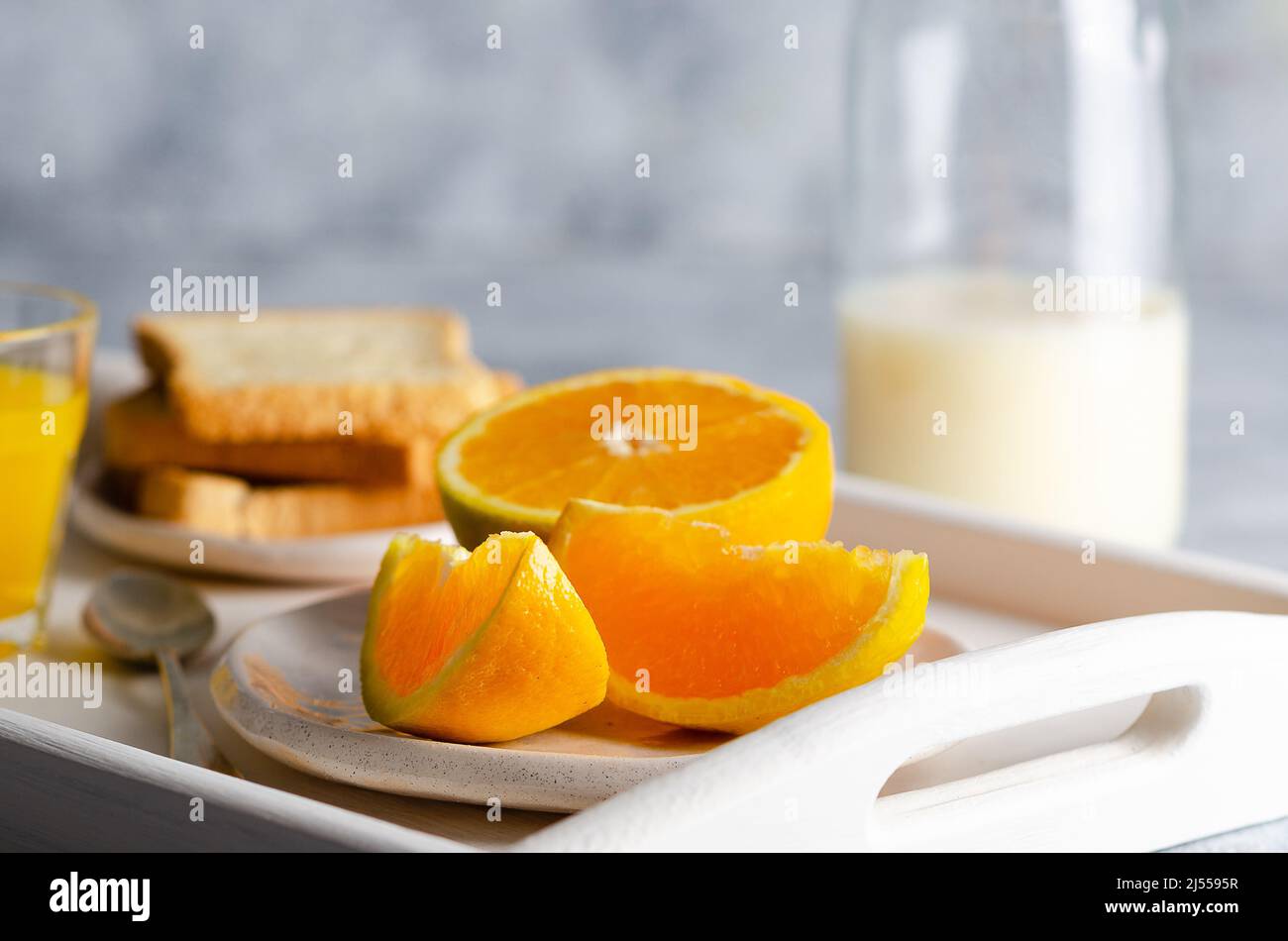 Una bottiglia di latte, un bicchiere di succo d'arancia e pezzi d'arancia, in un vassoio bianco e sfondo grigio. Foto Stock