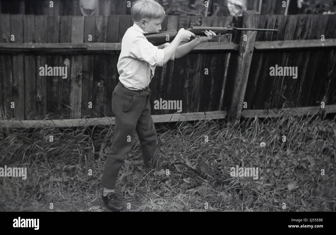 1960s, storico, all'esterno in un giardino posteriore, accanto ad una recinzione di legno, un ragazzo in piedi, a parte, tenendo un fucile ad aria in entrambe le mani in una posizione di tiro o tiro, eventualmente un modello Diana 27, Inghilterra, Regno Unito. Foto Stock
