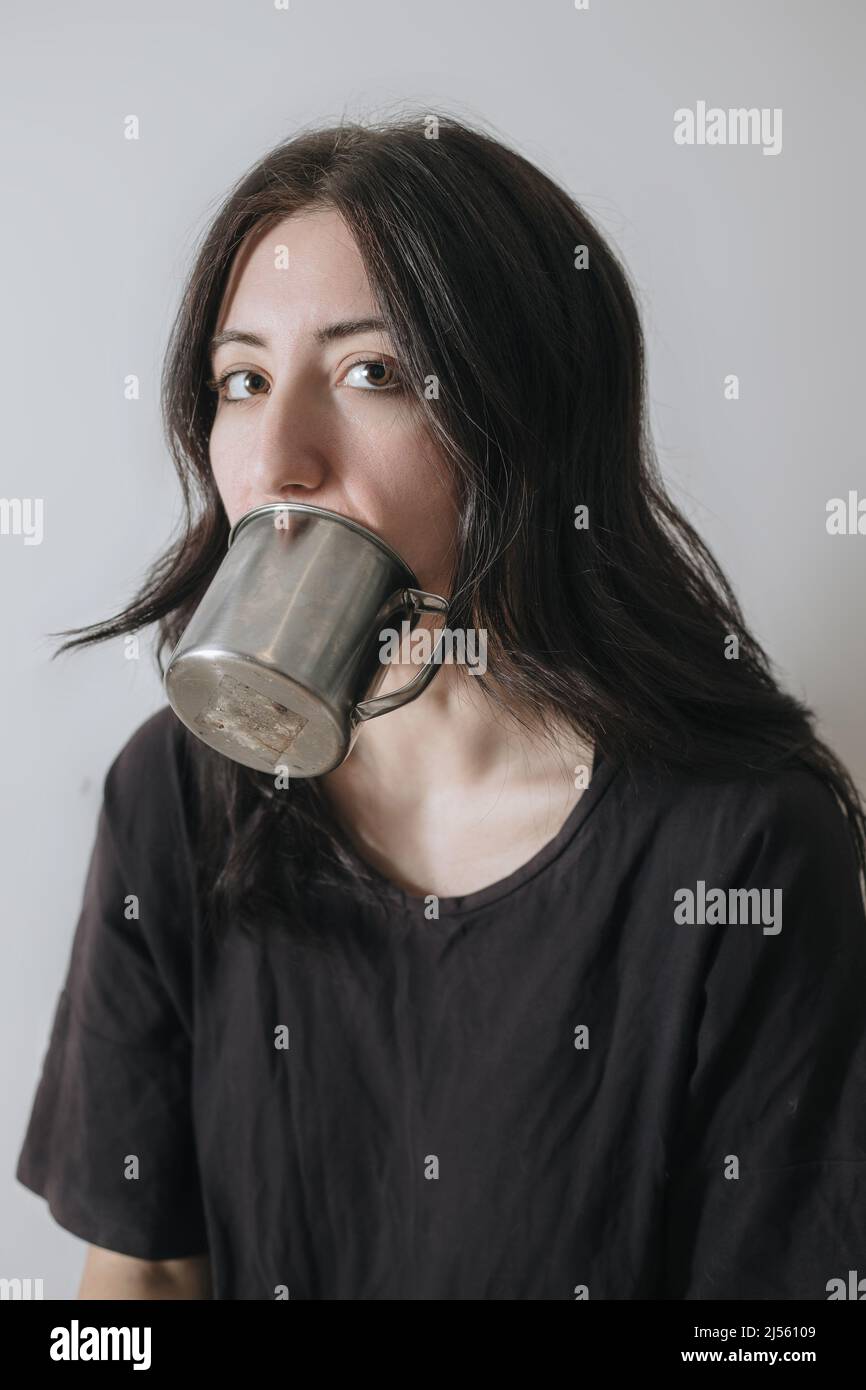 Giovane donna con una tazza di tè alla finestra durante la quarantena indossare maschera viso preoccupato per il nuovo stile di vita. Ansia, stress, salute mentale, crisi Foto Stock