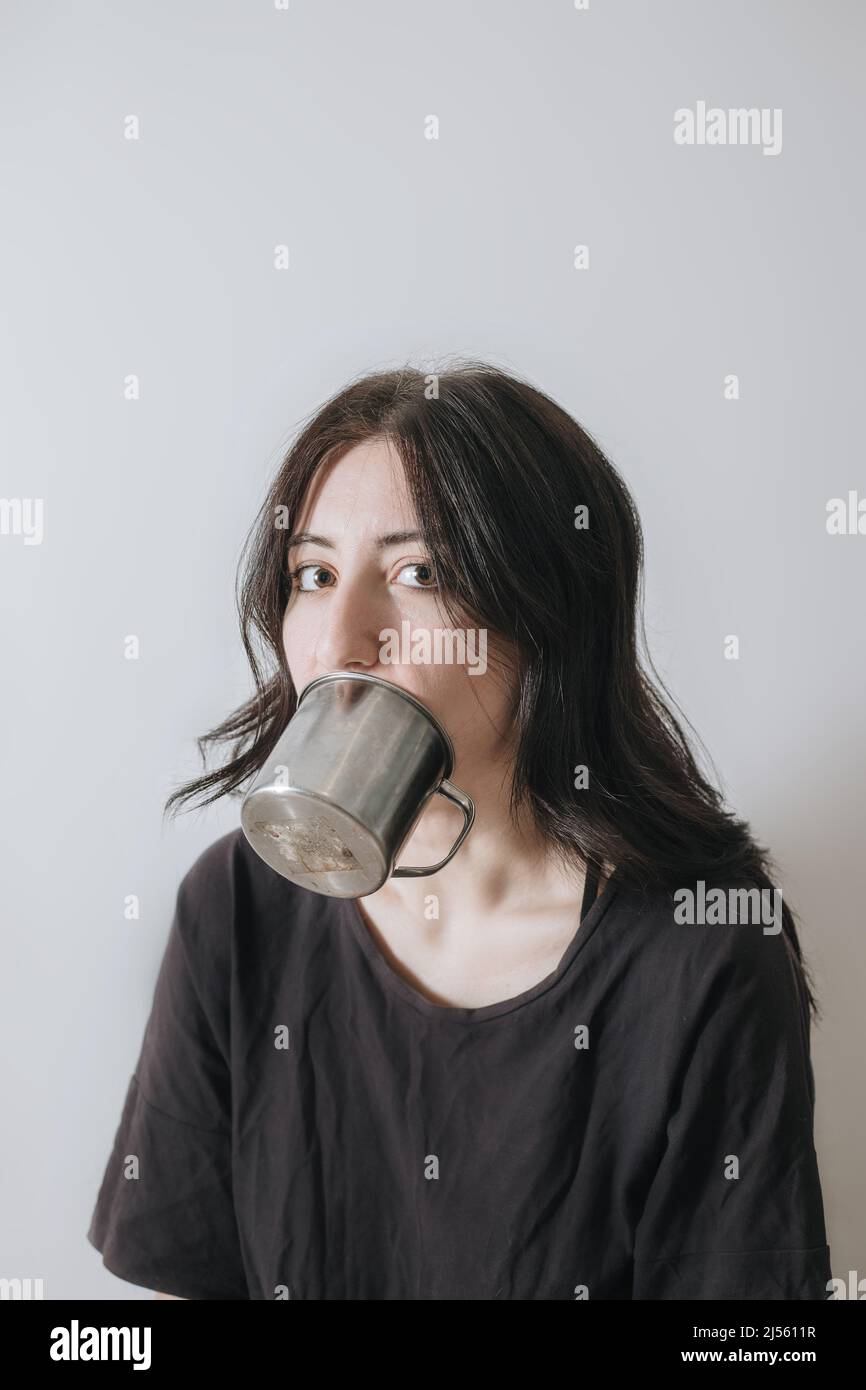 Giovane donna con una tazza di tè alla finestra durante la quarantena indossare maschera viso preoccupato per il nuovo stile di vita. Ansia, stress, salute mentale, crisi Foto Stock