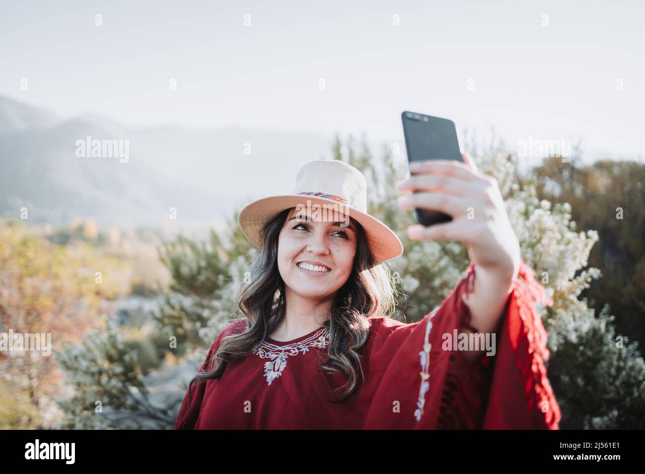 Sorridente giovane viaggiatore donna con un poncho rosso e un cappello, prendendo un selfie con il suo mobile in uno spazio naturale. Foto Stock