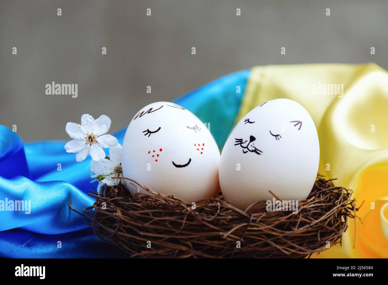 Uova di Pasqua bianche in un nido di vimini di legno con belle e graziose facce dipinte su uno sfondo satinato blu e giallo brillante della bandiera di Stato dell'Ucraina. Sfondo grigio. Con amore per il paese. Foto Stock